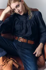 Camisa de denim azul oscuro con cuello mao, corte recto, mangas largas con puños abotonados, cierre frontal con botones a presión y detalle de volados en el cuello y la pechera.