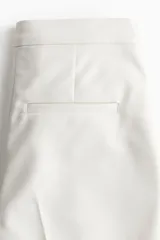Pantalón cigarette tobillero color blanco, con cintura estándar, cierre y pretina prolongada con corchete oculto. Piernas ajustadas con abertura corta en los bajos, bolsillos al bies y bolsillo de atrás decorativo. Pliegues adelante y atrás.
