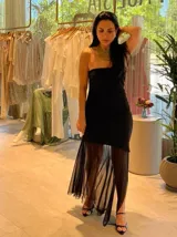 Vestido negro con escote strapless, corte ajustado al cuerpo y falda midi con abertura lateral y sobrefalda de tul transparente.
