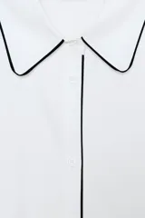 Camisa blanca de popelín con vivos negros a contraste en el cuello, el frente y los puños. Presenta un corte holgado y mangas largas con puños anchos.