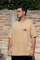 Remera color arena de manga corta con estampa en la espalda de un perro sentado junto a una tabla de surf.