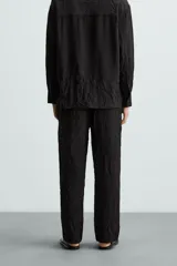 Pantalón negro de corte relajado (relaxed fit) con textura arrugada, tiro medio, bolsillos laterales y bolsillos traseros de vivo. Cierre frontal con cremallera y gancho metálico.