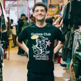 Remera negra de corte boxy con estampa de un personaje de historieta con gorra, dados y estrellas. Incluye texto "Gurises Club", "Uruguay", "Montevideo Cool Kids" y el logo de la marca Kaboa.
