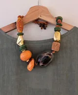 Collar con cuentas de diferentes formas, colores y materiales, incluyendo cuentas de resina naranja y verde, cuentas doradas, cuentas de madera y una cuenta grande con estampado animal print. El collar está ensartado en un cordón marrón.