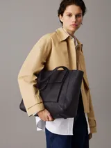 Cartera tipo shopper mediana de Calvin Klein, color negro, confeccionada en material textil con textura de lino tejido. Presenta asas fijas para el hombro, cierre de cremallera superior, bolsillo exterior frontal con cierre giratorio y herraje con monograma CK.