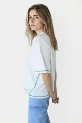 Remera blanca de corte holgado con cuello redondo y mangas cortas. Presenta un pequeño estampado de una flor amarilla en el centro del pecho y detalles de costura en azul en cuello, mangas y ruedo.