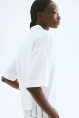 Camisa blanca de lino y viscosa, de corte holgado, con cuello clásico, botones frontales, mangas cortas y bajo redondeado.