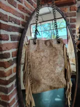 Bolso bandolera de cuero color dorado con textura craquelada y flecos largos en los laterales. Tiene cadena metálica dorada y herrajes dorados.