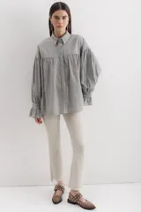 Camisa gris a rayas verticales blancas, de corte amplio, cuello alto con lazo y mangas largas abullonadas con puños elásticos.