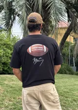 Remera de algodón negra con estampado de un balón de fútbol americano y la frase "Together, we are America". Incluye una firma en blanco debajo del balón.