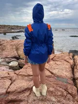 Piloto impermeable color azul eléctrico con detalles naranjas en los hombros, capucha integrada, cordón regulable en la cintura y mangas con volumen y puños elastizados.