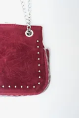 Cartera de mano o clutch color rojo oscuro (bordó) con textura de gamuza o terciopelo. Presenta tachas metálicas plateadas a lo largo del borde inferior y lateral, y un cierre perimetral plateado. Incluye una correa de hombro desmontable compuesta por una tira de material a tono y cadena metálica plateada.