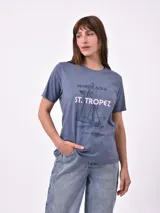 Remera blanca de algodón con cuello redondo y manga corta. Presenta un pequeño bordado en el pecho izquierdo con una figura que parece ser una persona acostada en una tabla de surf o flotador.