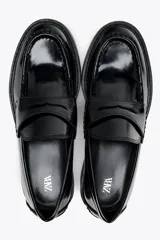 Zapato tipo mocasín de charol negro, con detalle de antifaz en el empeine y punta redonda.