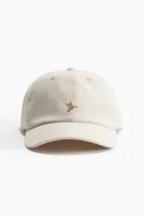 Gorra color crema de sarga de algodón con diseño bordado de un pájaro en el frente. Tiene ojales bordados en la parte superior, trabilla ajustable con hebilla de metal atrás y cinta antitranspirante de algodón.