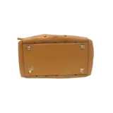 Cartera bombonera color marrón claro, confeccionada en cuero con apliques de lona revestida con el monograma de la marca y pequeñas tachas decorativas. Tiene correa para llevar al hombro, bolsillo interior deslizante y cierre con cordones ajustables que le dan un efecto fruncido.