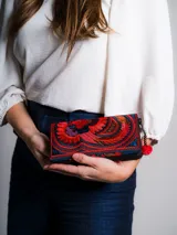 Billetera de tela bordada con motivos florales y de aves en tonos rojo, azul y morado. Cierre con cremallera y detalle de borla roja y cuentas de madera. Confeccionada artesanalmente en Tailandia.