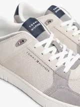 Zapatillas de corte bajo inspiradas en modelos retro de baloncesto, con exterior de ante y paneles de malla en tonos grises y beige. Presentan suela de goma blanca gruesa, cierre de cordones y detalles distintivos de la marca Tommy Hilfiger en el lateral, lengüeta y talón.