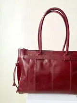 Bolso grande de cuero marrón con textura tipo cocodrilo, estructura rígida y asas para llevar al hombro. Tiene una tira con tachas y hebillas en cada extremo, también lleva lazos en los costados.