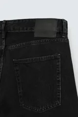 Jeans de denim de algodón, color negro lavado, con tiro medio, corte ajustado en cadera y muslos, y pierna que se estrecha hacia el tobillo. Presenta cinco bolsillos y cierre frontal con cremallera y botón.