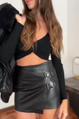 Minifalda negra de cuero ovino, de tiro bajo, con corte irregular y detalle de hebillas metálicas en la cintura.