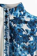 Vestido corto camisero blanco con estampado floral azul, cuello mao, botonadura frontal y cinturón fino para atar extraíble. Mangas globo de corte raglán, puños abotonados y costura fruncida bajo la cadera.