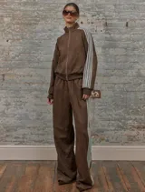 Conjunto deportivo marrón de dos piezas: campera con cremallera, cuello alto, mangas largas con puño y detalles de rayas blancas en mangas y laterales del pantalón. Pantalón recto de cintura alta con cordón ajustable.