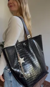 Bolso tote negro con textura símil cuero de cocodrilo, doble asa de hombro marrón y cierre con botón a presión. Incluye un charm colgante con una estrella blanca y cadenas plateadas.