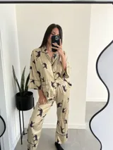 Pijama de dos piezas color beige con estampado de tigres negros y ribetes negros. Incluye camisa de manga larga con cuello y botones, y pantalón largo de corte recto.