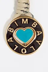 Llavero con mosquetón y anilla metálica plateada grabada con el logo de Bimba y Lola. Presenta un cordón trenzado verde con detalles en blanco que sujeta un dije circular con un corazón rojo esmaltado en el centro y el nombre de la marca calado alrededor.