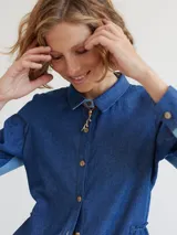 Camisa celeste de algodón con cuello clásico, cierre frontal con botones y mangas largas. Presenta volados en el frente y corte evasé.