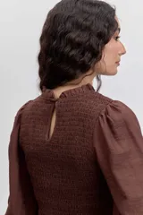 Blusa color marrón con cuerpo fruncido tipo nido de abeja, cuello alto con volados y mangas largas abullonadas con puños elásticos terminados en volados.