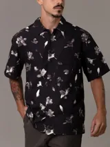 Camisa de manga corta con estampado de flores y hojas en blanco y gris sobre fondo negro. Tiene cuello clásico, cierre con botones y bolsillo de parche en el pecho.