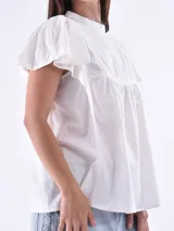 Blusa blanca de manga corta con volados, cuello alto y detalles de encaje o bordado vertical en el frente. Presenta un corte holgado con frunce bajo el busto.