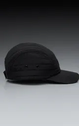 Gorra negra de estilo 5 paneles, con tejido acolchado o con costuras horizontales y visera plana. Presenta un pequeño parche cuadrado negro con un logo blanco bordado en forma de 'X' o lazo.