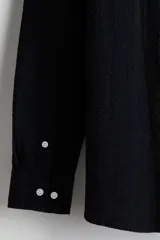 Camisa negra de sirsaca de algodón con cuello inglés, tapeta clásica, bolsillo superior de parche y canesú con pliegue de caja en la espalda. Mangas largas con tapeta de manga y botón, y cierre ajustable de botón en los puños. Bajo redondeado y corte holgado.
