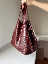 Cartera tipo tote bag color bordó, confeccionada en cuero con textura granulada. Tiene doble asa corta de mano y asa larga extraíble con diseño de tachas para usar como bandolera.