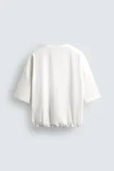 Camiseta blanca de algodón, corte holgado, cuello redondo, manga corta y bajo con elástico interior.
