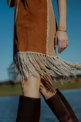 Vestido corto marrón de gamuza sintética, sin mangas, con escote en V y detalle de flecos en el ruedo. Presenta apliques bordados en el frente y laterales.