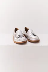 Mocasín de cuero color marrón claro, con detalle de flecos y borlas en la parte delantera. Diseñado en Uruguay y fabricado en Brasil.