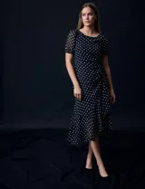 Vestido midi de gasa color negro con lunares blancos, con mangas cortas, escote redondo y ruedo asimétrico. Presenta un frunce lateral con lazo de ajuste.