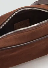 Bolso pequeño de piel bovino color marrón oscuro, con acabado en serraje (gamuza). Presenta un forro interior de algodón y cierre principal de cremallera. Incluye un asa larga ajustable.