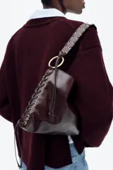 Bolso de hombro marrón oscuro con acabado brillante, asa trenzada y ojales metálicos en los laterales.