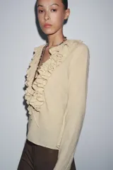 Camisa color beige de algodón con escote en V y volantes. Tiene mangas largas con botón en los puños y cierre con lazos en el frente.