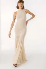 Vestido largo color beige, con cuello alto y sin mangas. La parte superior es ajustada al cuerpo y la falda es amplia y fluida, con paneles traslúcidos.
