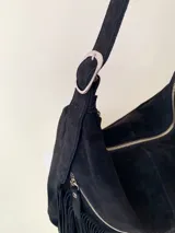 Bolso tipo hobo color tostado, hecho de cuero con flecos largos en el frente y dos bolsillos con cierre. Tiene una manija regulable con hebillas metálicas.