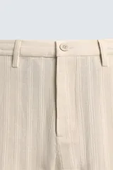 Pantalón chino de corte relajado, color beige claro, con textura de rayas verticales sutiles. Presenta cintura ajustable con cordón interior, bolsillos delanteros y bolsillos traseros de vivo.