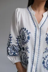 Camisa blanca de manga larga con elástico en puños, cuello mao con botones en la parte delantera y bordados florales azules en el frente y las mangas.