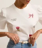 Remera blanca de algodón con cuello redondo y mangas cortas abullonadas. Presenta estampados de un lazo, una flor, un corazón y frases caligráficas.