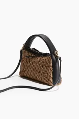 Cartera pequeña con cuerpo de paja trenzada color marrón claro y laterales de cuero sintético negro. Tiene asa corta de mano y correa larga extraíble para usar cruzada.